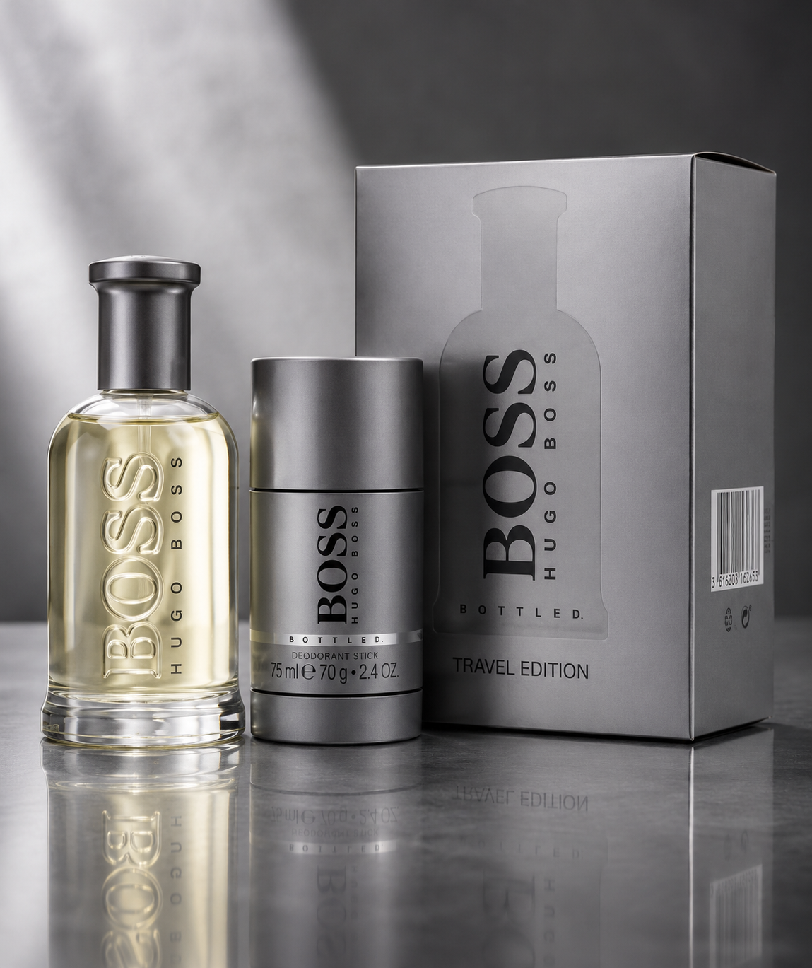 BOSS Hugo Boss Bottled Travel Edition fragrance set featuring a clear glass bottle with pale yellow liquid, a metallic silver cylindrical deodorant stick, and a large silver-gray product box on a reflective gray surface.